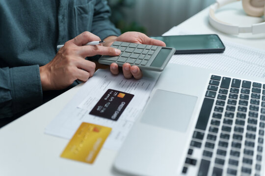 Man using a calculator beside credit cards, financial documents, laptop and smartphone at a desk, representing budgeting, debt management, expense tracking, payments and personal finance planning.