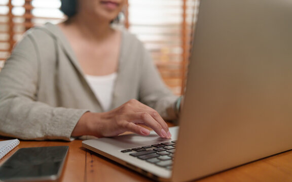 Asain Woman types on a laptop at a home desk with a smartphone and notebook nearby, representing remote work, freelance writing, online business, digital communication and modern work productivity.