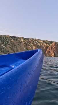 Blue kayak bow glides across calm sea water toward towering rocky mountains at golden hour. First-person perspective steady shot.