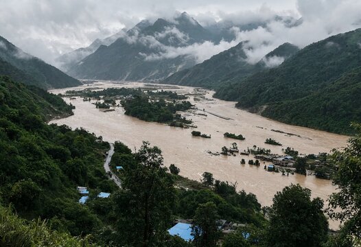 A wide river with brown flood water flowing through a mountainous valley landscape during a cloudy day