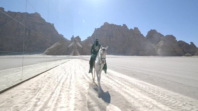 Cinematic shot of Saudi female equestrian in green cloak and veil proudly raising sword while riding white horse in front of Maraya in AlUla