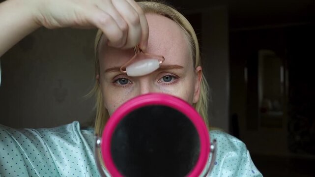 Concerned woman uses a quartz facial roller to massage forehead wrinkles and glabellar lines. She looks in the mirror, seeking anti aging skin care alternatives to botox.