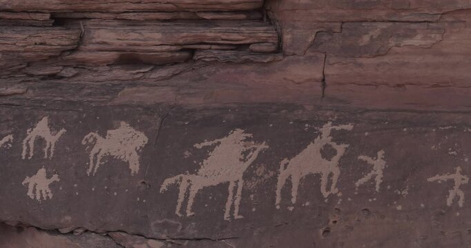 Cinematic close-up of ancient petroglyphs and rock art depicting camel caravans and armed horsemen on sandstone cliff in AlUla