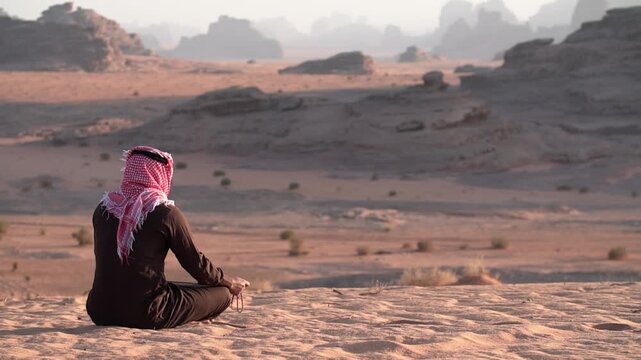 Cinematic profile shot of Saudi man in warm winter thobe and shemagh holding rosary beads while contemplating sunset over Hisma sand dunes in NEOM Saudi Arabia