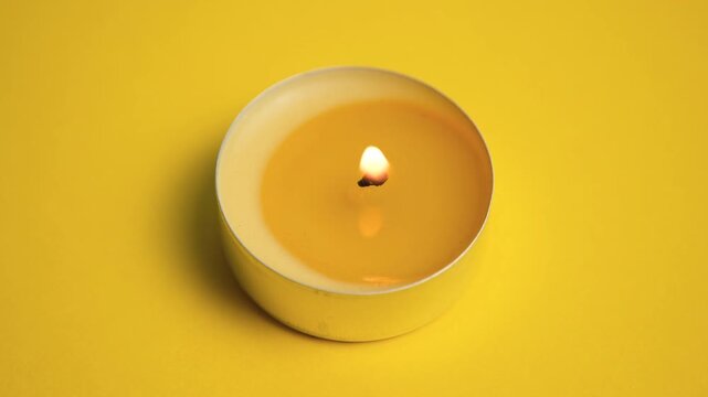 Single burning yellow tealight candle with warm glowing flame on yellow background, minimalist close-up composition.