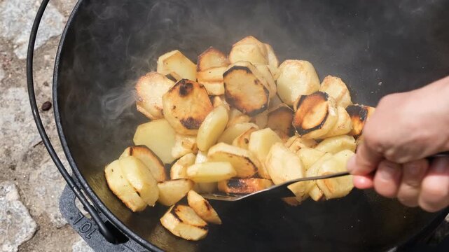 Sliced potatoes frying and turning golden brown in a large black cast iron cauldron while being stirred with a spatula.