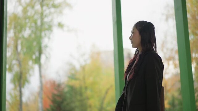 Woman in black blazer and white pants walks through outdoor area with green structural elements holding phone. Woman carries brown bag while moving past trees showing autumn foliage