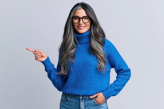 Mature woman with long grey hair wearing blue sweater and glasses pointing to side