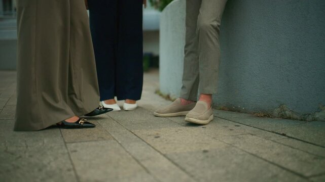 Three business people stand together, engaged in a conversation outdoors. Their legs and feet are visible as they talk during a casual outdoor meeting.