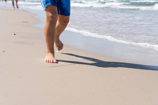 Barefoot Child Walking On Sandy Beach By Sea During Summer Vacation And Coastal Travel