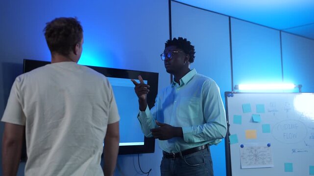 Diverse male software developers discussing codes on a large screen in modern IT office. African American and Caucasian programmers analyzing cybersecurity data. Corporate technology teamwork concept