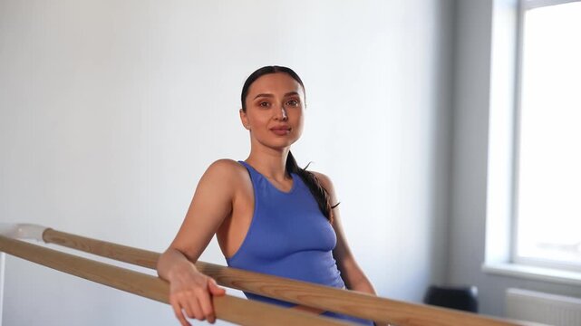 Portrait of attractive fitness woman in activewear looking with warm radiant smile at gymnastic barre, expressing natural beauty, confidence, happiness and wellness while posing indoors.