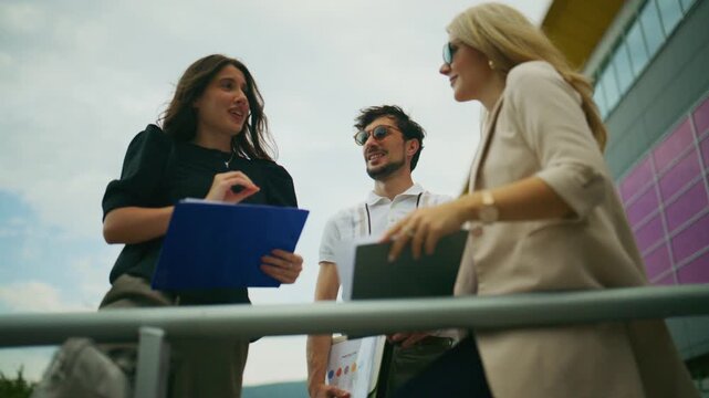 Three business professionals discuss work outdoors with documents. The colleagues share a positive and collaborative meeting.
