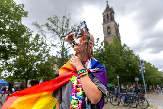 Masked therian girl in colorful rainbow cape poses during CSD celebration in Magdeburg, Germany, with cathedral behind. Scene reflects youth subculture, fandom, identity, pride, creativity, community