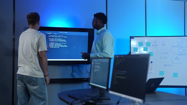 Diverse male software developers discussing codes on a large screen in modern IT office. African American and Caucasian programmers analyzing cybersecurity data. Corporate technology teamwork concept
