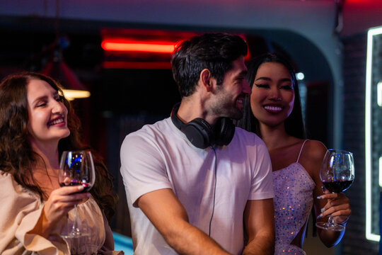 A smiling bearded DJ in a white t-shirt flanked by two young women holding red wine cues a track while the two women dance on either side at the DJ booth of a neon-lit nightclub.