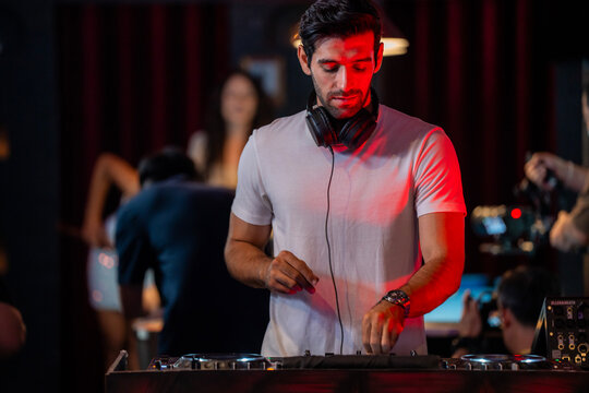 A wide view of a male DJ standing behind turntables and a mixer in a busy nightclub concentrates on the turntables with the curtain glowing behind in a warm candid medium shot.