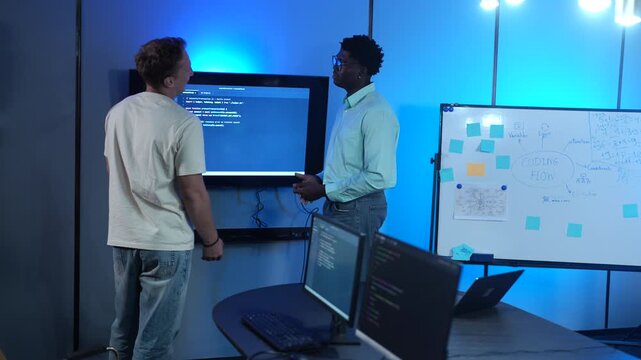 Diverse male software developers discussing codes on a large screen in modern IT office. African American and Caucasian programmers analyzing cybersecurity data. Corporate technology teamwork concept