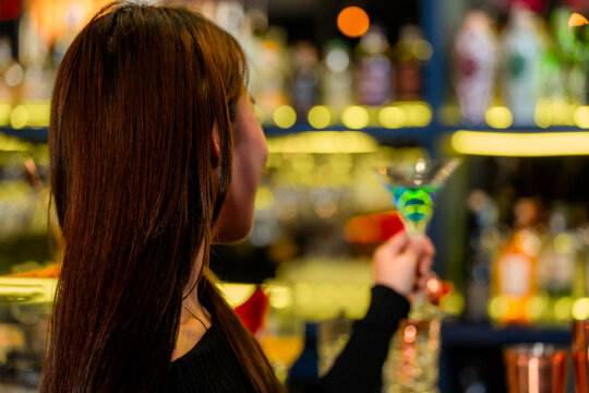 A beautiful young Asian woman in a black off-shoulder top holding a bright blue cocktail brings the blue martini slowly toward her lips at the polished counter of a cocktail bar.