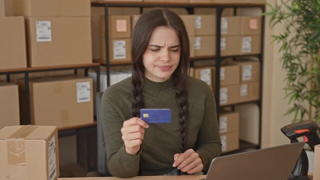 Woman holding creditcard in hand, frowning at laptop amid packed boxes in a building; payment problem frustration.