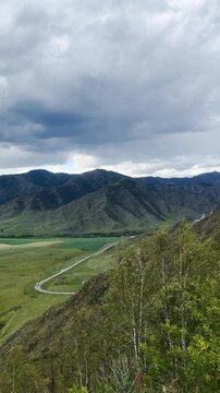Moody mountain valley with heavy clouds, sweeping green grassland, forested slopes, winding road, layered ridges, pale river channel, and elevated hillside lookout
