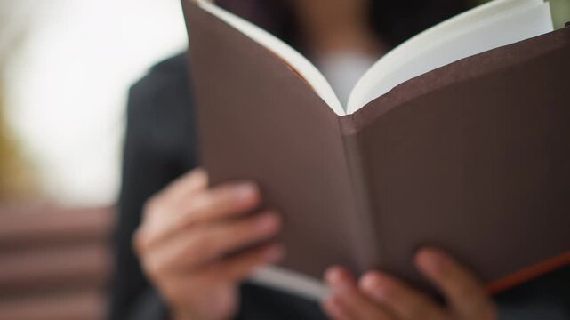 Close up of woman holding and reading open book with white pages. woman wears dark clothing focusing on hands gripping brown hardcover notebook. Soft natural light illuminates scene creating