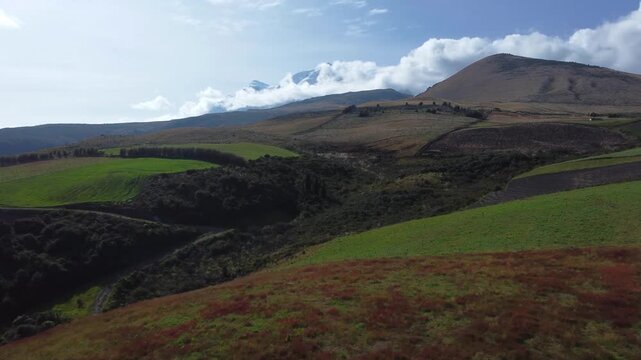 Aerial drone shot of the snow capped Cayambe volcano rising behind rolling paramo hills covered in red and green highland vegetation in the Ecuadorian Andes.