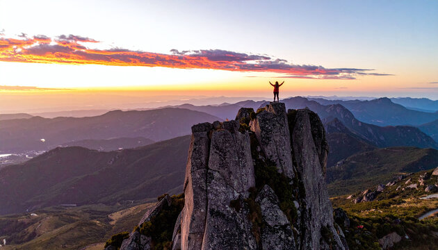 A triumphant hiker stands atop a dramatic mountain peak with arms raised at vibrant sunset. An inspiring aerial view evoking freedom, achievement, and awe for travel concepts.