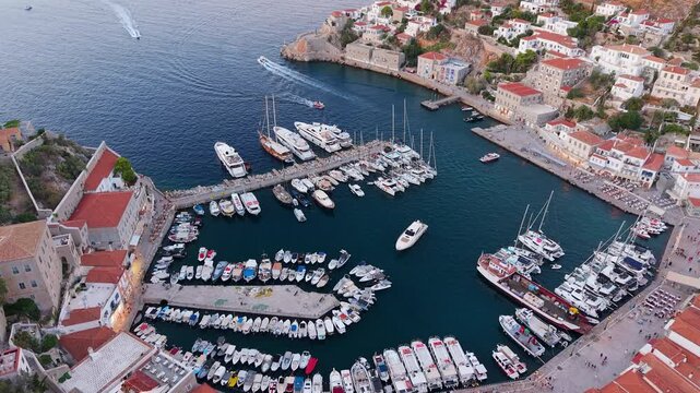 Drone flies over Hydra's horseshoe harbor from above at dusk. The iconic marina packed with boats, the waterfront promenade and historic mansions form a stunning geometric pattern.