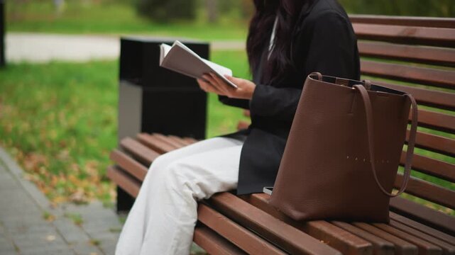 Close up of woman sitting on wooden bench in outdoor park reading book while wearing white pants and black shoes. brown leather bag rests beside her as woman remains seated throughout scene