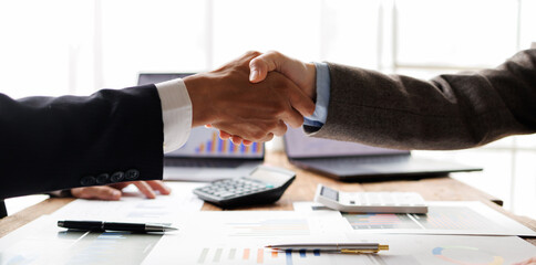 Two businesspeople shaking hands over a desk filled with documents, laptops, and calculators, symbolizing a successful partnership, agreement, and closing of a deal © groviapoto