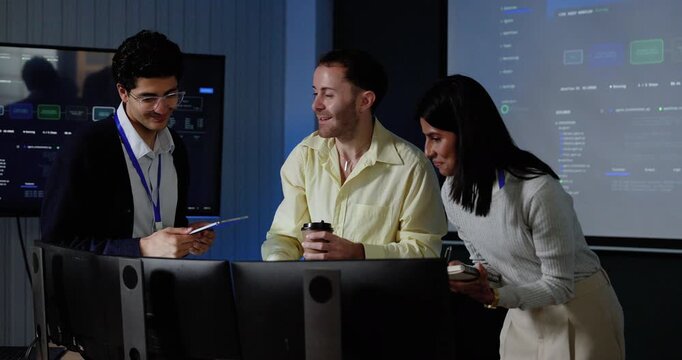 Young Asian man, adult Caucasian man and young Asian woman lean toward computer monitors, exchange gestures and review software dashboard during technology discussion and digital system analysis.