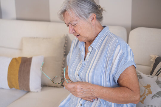 Senior adult woman feeling unwell with stomach pain and holding glasses, sitting on a sofa at home, experiencing discomfort, illness, and digestive issues