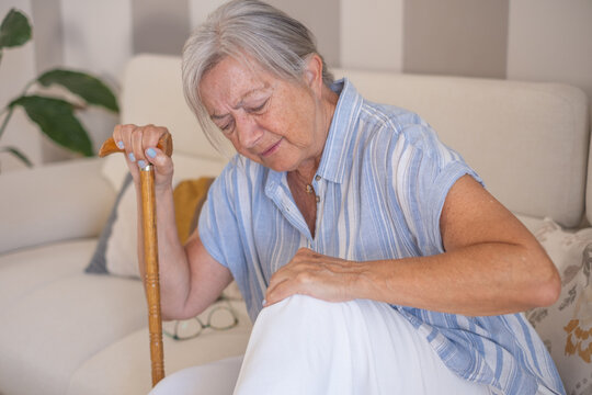 Senior woman experiencing sharp knee pain while sitting at home, holding a walking stick and needing assistance with health and mobility issues