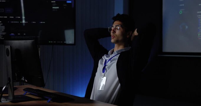 Young Asian man stretches at computer workstation, covers face and adjusts glasses while pausing from software work, conveying office fatigue, workplace stress, system analysis, digital workflow.
