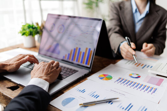 Business professionals analyzing financial data using a laptop and various charts on paper, collaborating on market strategy and corporate growth planning in an office setting