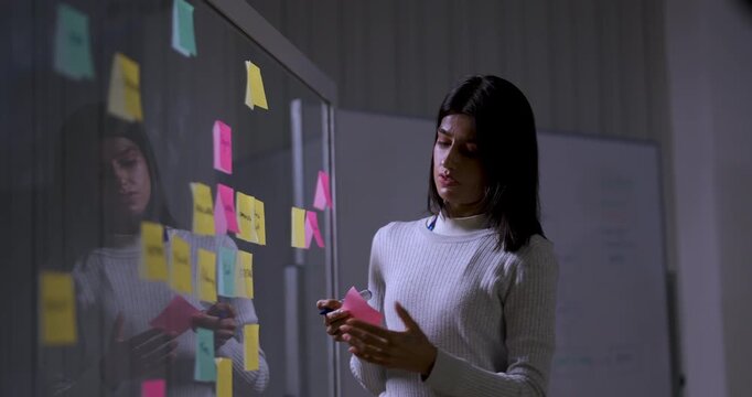 Adult female technology professional arranges sticky notes on glass board, writing and pointing while planning software project workflow during business strategy session in modern corporate office.