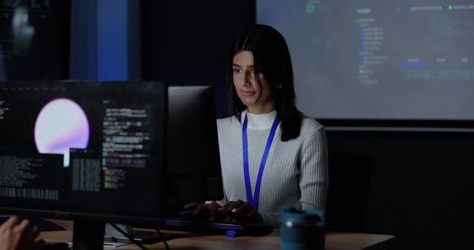 Young adult woman typing at computer workstation with multiple monitors and projected software interface, reviewing code during system analysis, cybersecurity workflow, digital project planning.