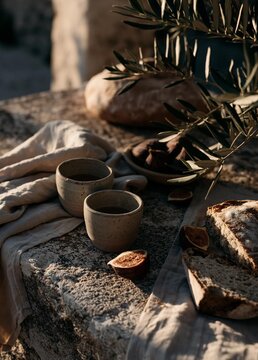 Stone table with two small bowls of coffee and a loaf of bread on it. the table is covered with a beige cloth and there is a branch of an olive tree on the right side of the table.