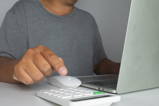 Close up of a man working on a laptop, using a white calculator and mouse at a desk. Concept of accounting, financial planning, calculating budget, and online business management.