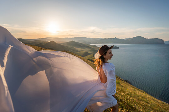 Woman hat dress, elegant female stands on a grassy mountain cliff overlooking a serene sea during a beautiful golden sunset