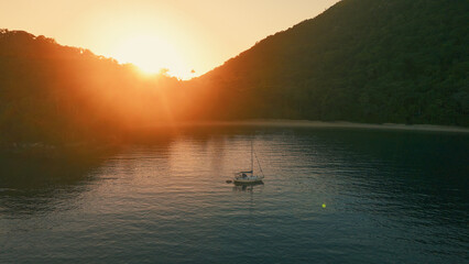 Obraz premium Aerial view of the sailing boat anchored in the calm lagoon with sun rising and shining from the mountains