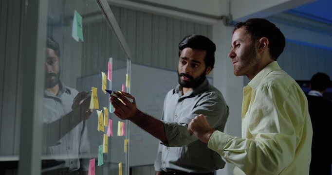 Two adult male technology professionals review sticky notes on glass board, gesture toward project tasks and exchange ideas while discussing software workflow during collaborative strategy session.