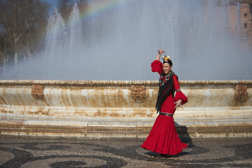 An Andalusian woman, wearing a red flamenco dress and a black shawl, with flowers in her hair, dancing flamenco next to a beautiful fountain in Seville’s Spain Square. A concept of dance and art. © Manuel