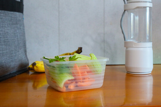 Fresh Chopped Vegetables Rest Inside Translucent Plastic Container On Polished Wooden Counter, With Celery, Parsley, Carrot Pieces, And Portable Blender Cup Creating Simple Healthy Meal