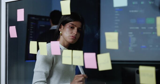 Adult female technology professional writes on sticky notes and places them on glass board, organizing software project ideas and workflow tasks during business strategy planning in modern office.