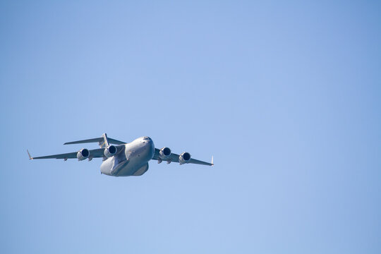 C-17 Globemaster III military transport aircraft in flight, viewed from below against a blue sky with a white cloud below the aircraft, showing its four jet engines and distinctive high-wing design.