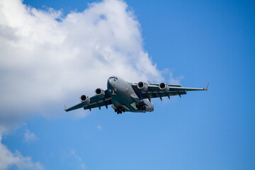 Naklejka premium C-17 Globemaster III military transport aircraft in flight, viewed from below against a blue sky with a white cloud below the aircraft, showing its four jet engines and distinctive high-wing design.