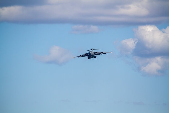 C-17 Globemaster III military transport aircraft in flight, viewed from below against a blue sky with a white cloud below the aircraft, showing its four jet engines and distinctive high-wing design.