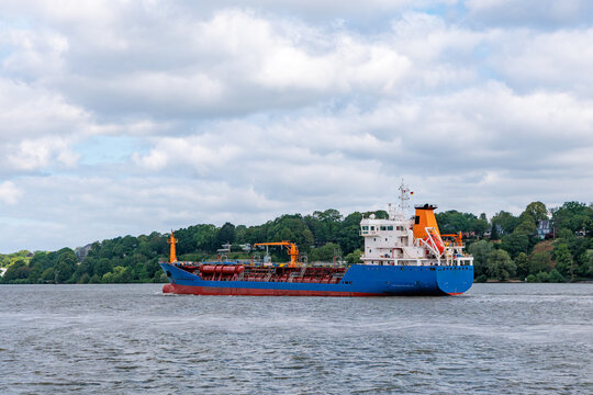 Blue and red chemical tanker ship navigating on river along green riverbank under cloudy sky, maritime freight transport and energy logistics concept